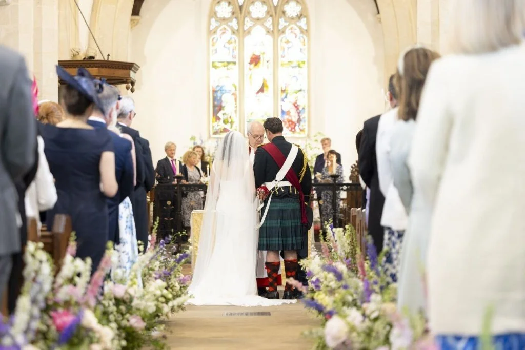 A wedding ceremony is taking place inside a church, with a bride and groom exchanging vows at the altar, surrounded by guests and floral decorations.
