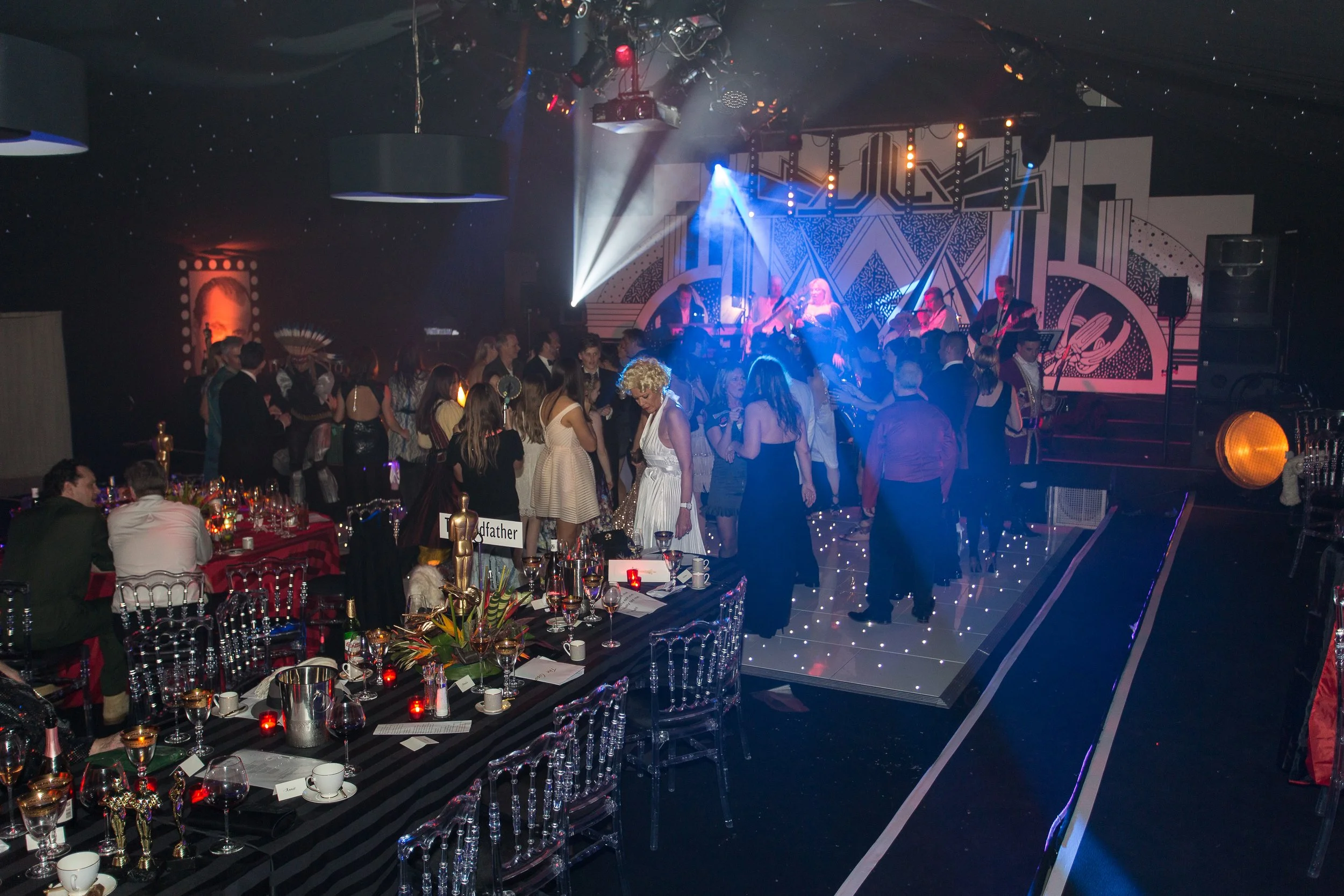 A lively evening event with a stage featuring live musicians and a crowd of guests dancing and socializing under dramatic stage lighting.