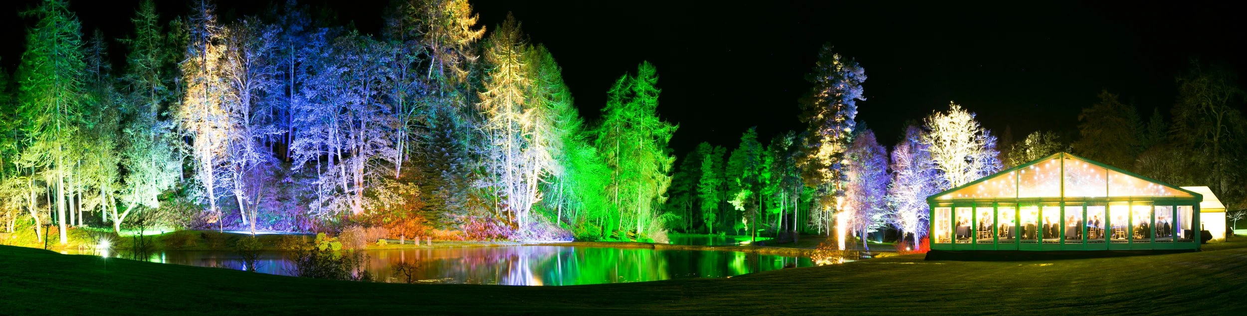 Colorfully lit trees surrounding a lake at night with a brightly lit marquee tent on the right side.