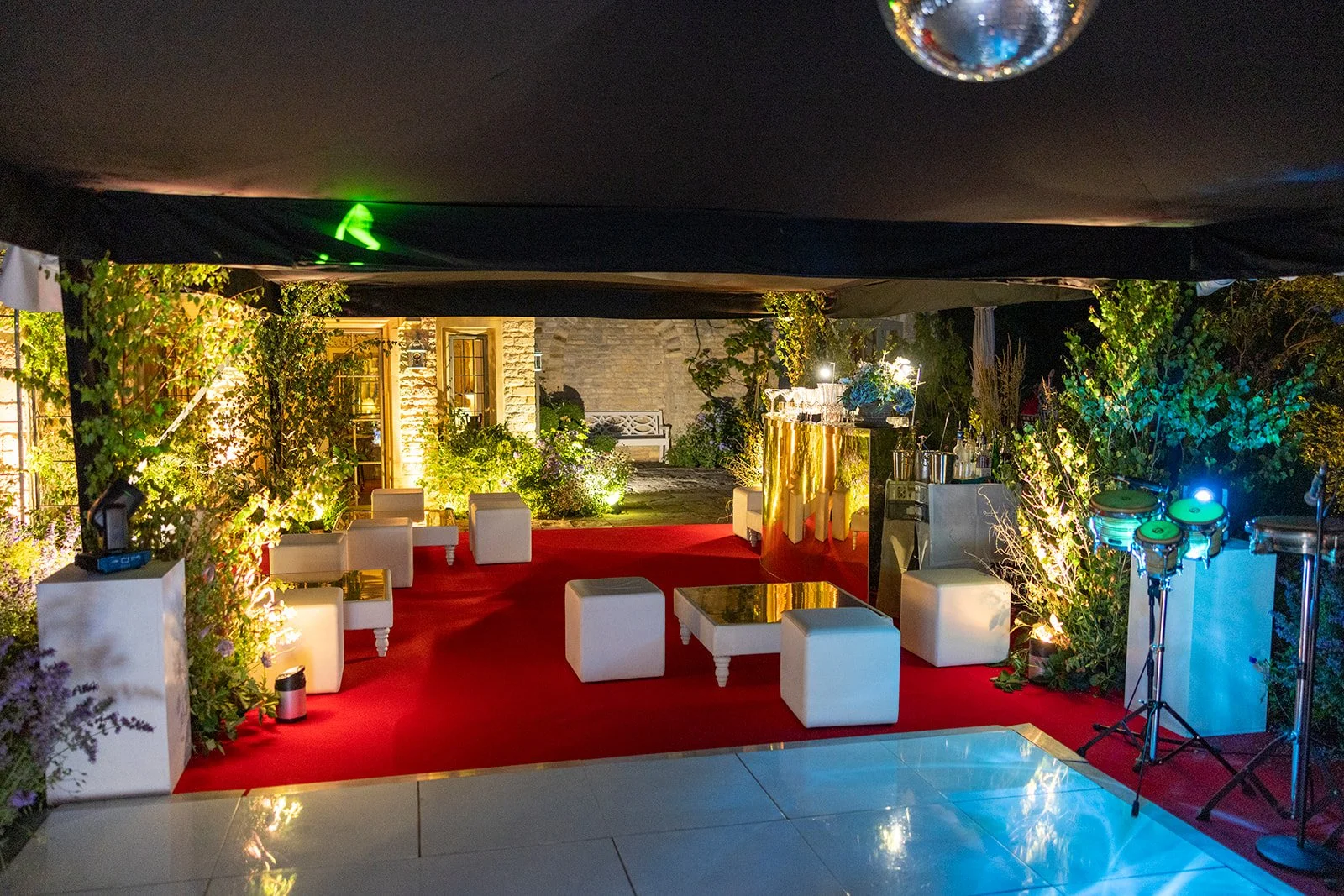 Nighttime outdoor event setup with white seating, decorative plants, and colorful lighting, including a red carpet, a bar with drinks, and percussion instruments on a stand.