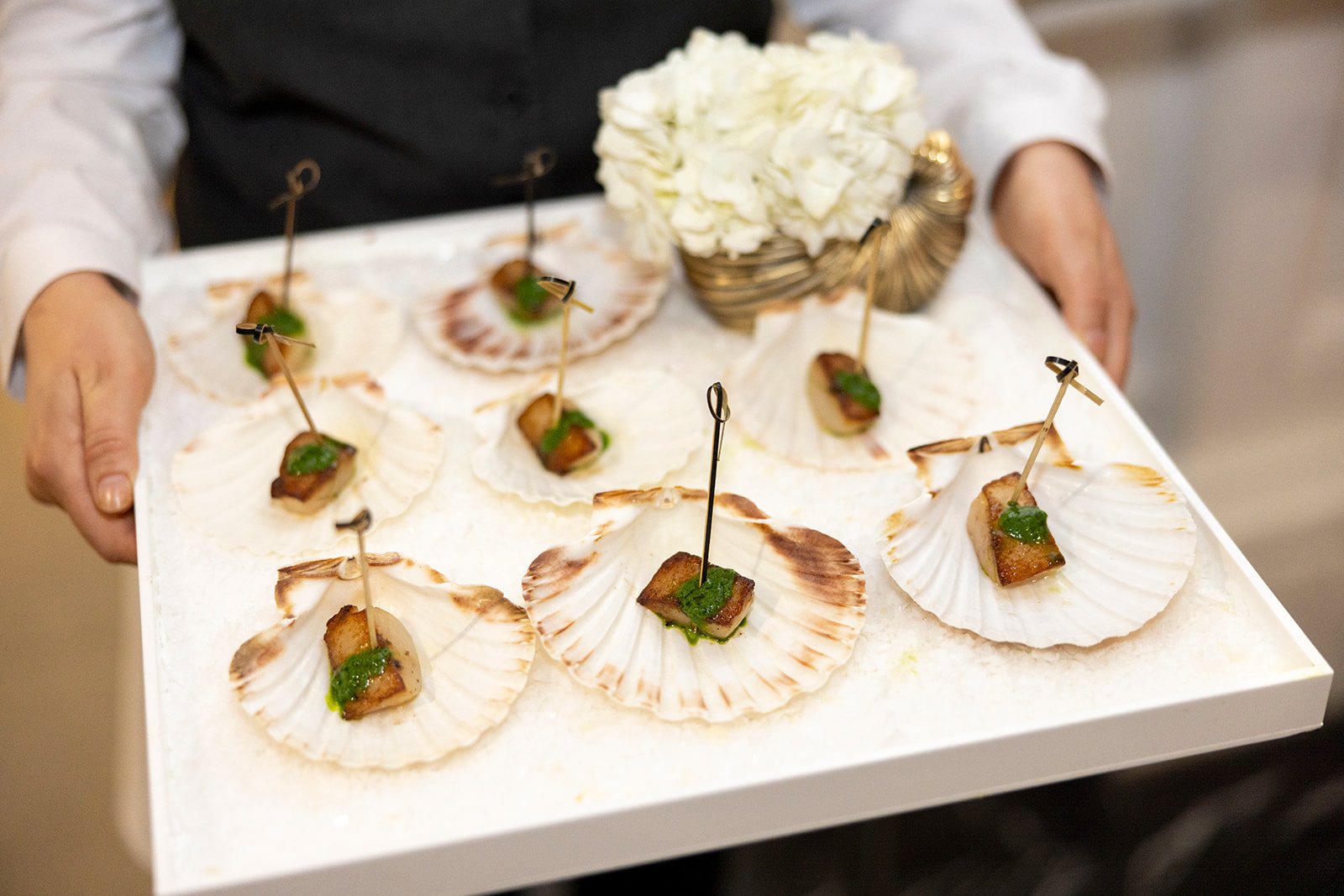 A person serving a tray of scallops in shells topped with a small piece of cooked meat and green sauce, garnished with a small skewer.