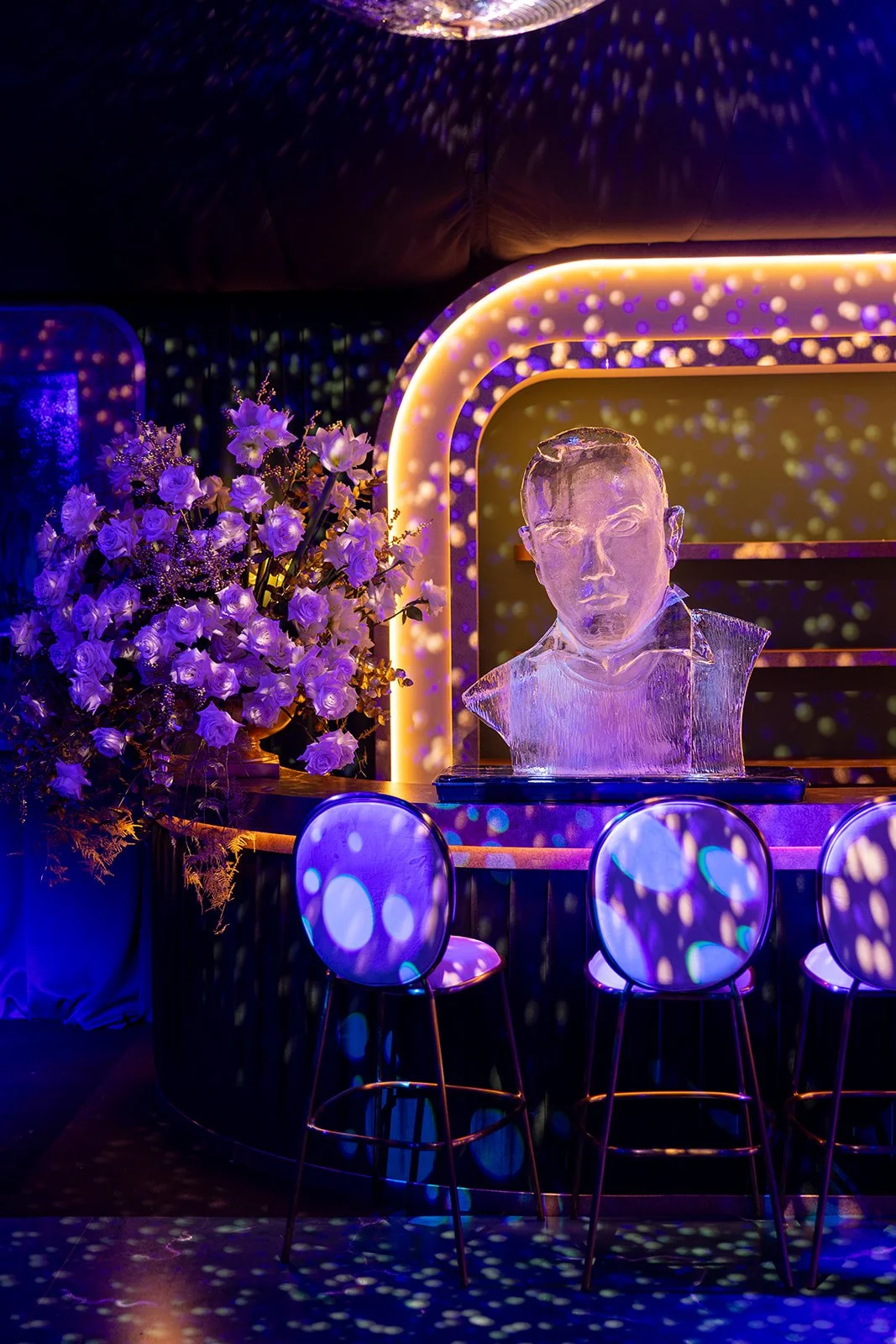 Ice sculpture bust of a man on a bar with purple and blue lighting, flowers, and circular light patterns.