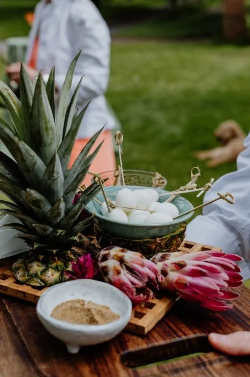 Pineapple, dragon fruit, and a bowl of eggs on a wooden surface outdoors with people and a dog in the background.