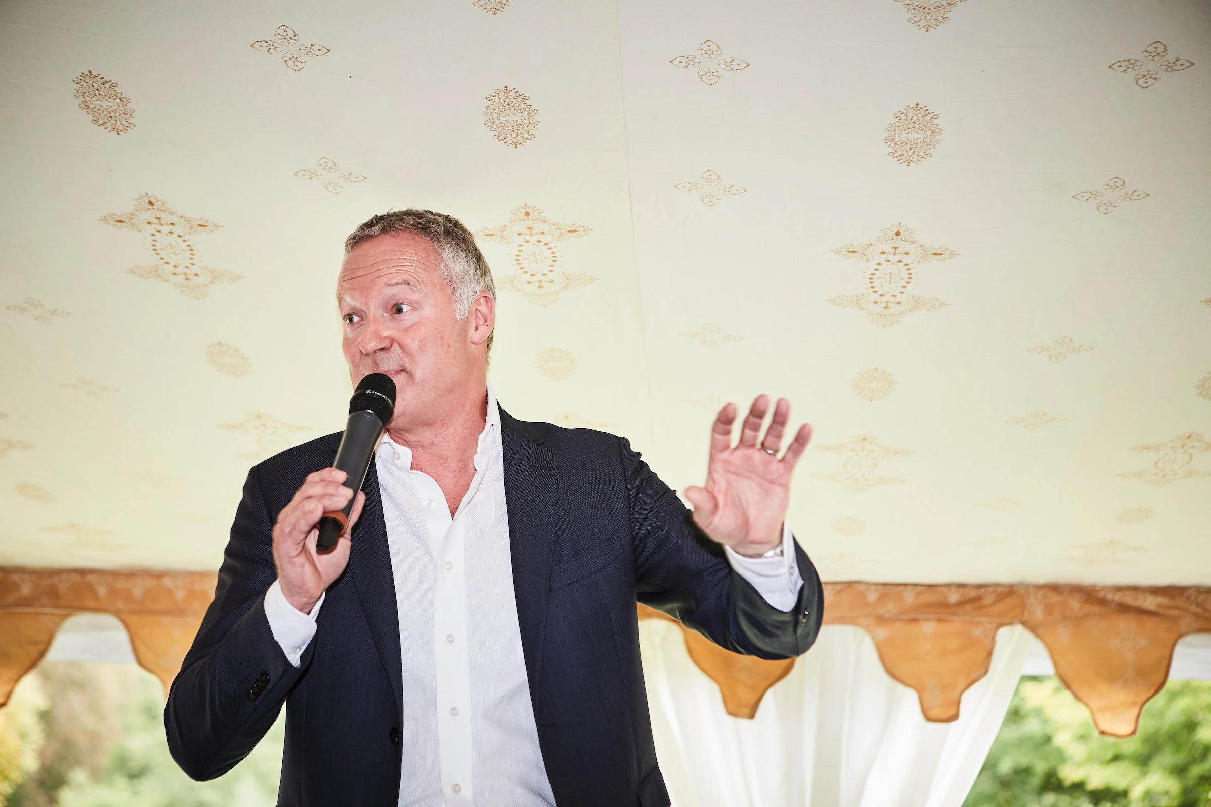 A man in a dark suit and white shirt speaking into a microphone, gesturing with his right hand, standing in front of a decorative wooden backdrop with a cream-colored wall and patterned ceiling.