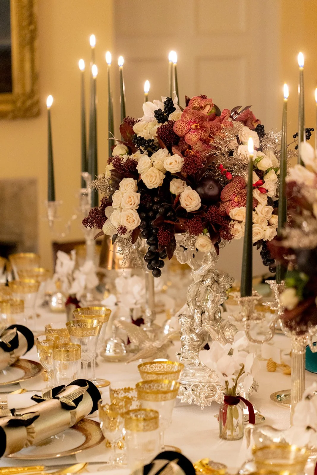 Elegant table centerpiece with white roses, orchids, black grapes, and greenery, surrounded by gold-rimmed glassware, ornate silver candlesticks with lit candles, and black and gold tableware, set in a luxurious dining setting.