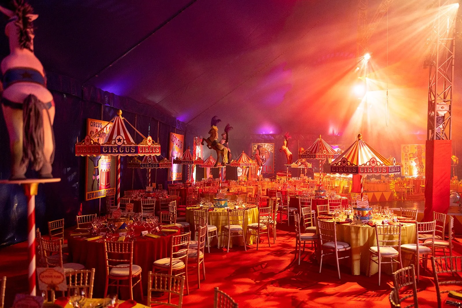 Interior of a circus-themed event tent with tables, chairs, and circus decorations, including figures of acrobats and circus tents, illuminated with warm orange and purple lighting.
