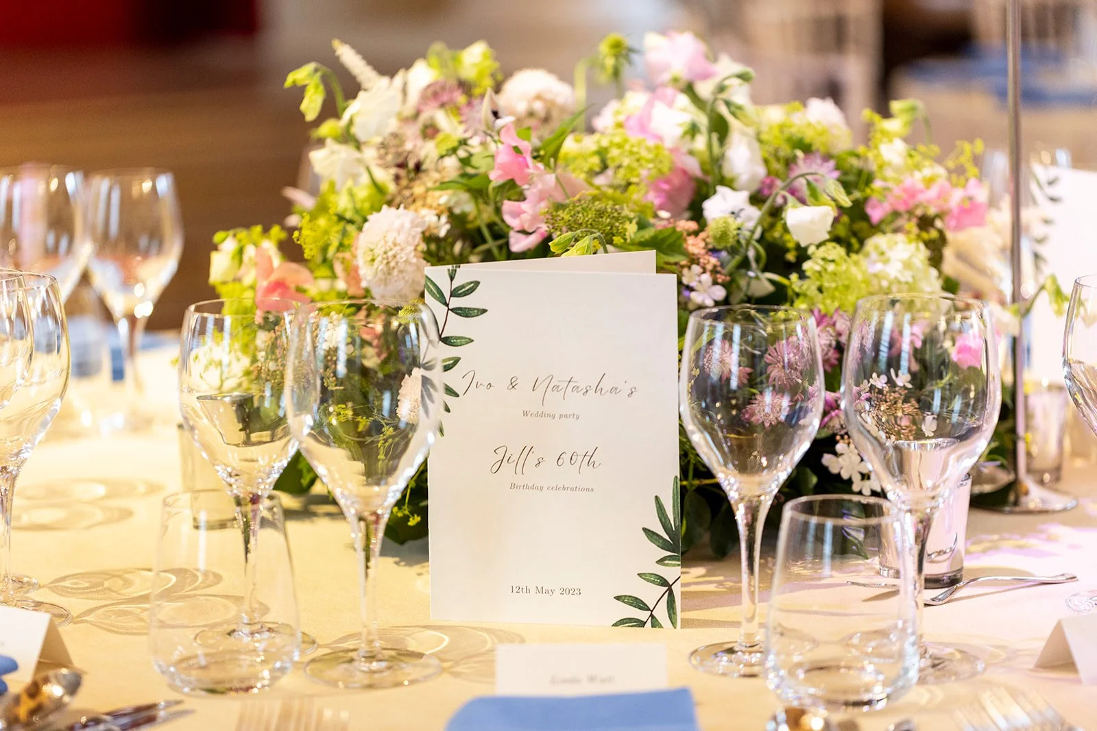 A wedding celebration table decorated with a large floral centerpiece featuring pink, white, and green flowers, multiple wine glasses, and a wedding program card indicating the celebration of Jill's 60th birthday on May 12, 2023.