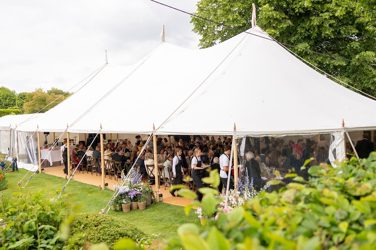 Large white event tent filled with people attending a gathering or celebration outdoors, surrounded by greenery and flowers.