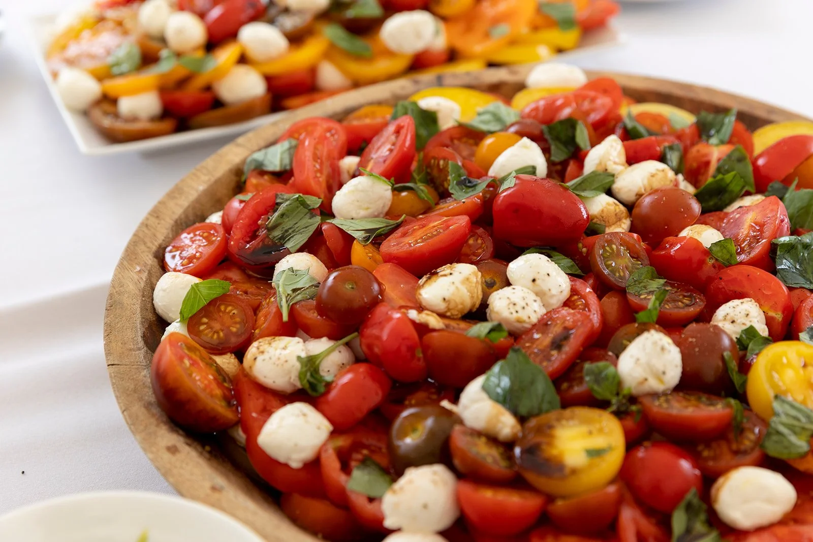 A wooden bowl filled with a cherry tomato salad with small mozzarella balls, fresh basil, and black pepper, with a plate of caprese salad in the background.