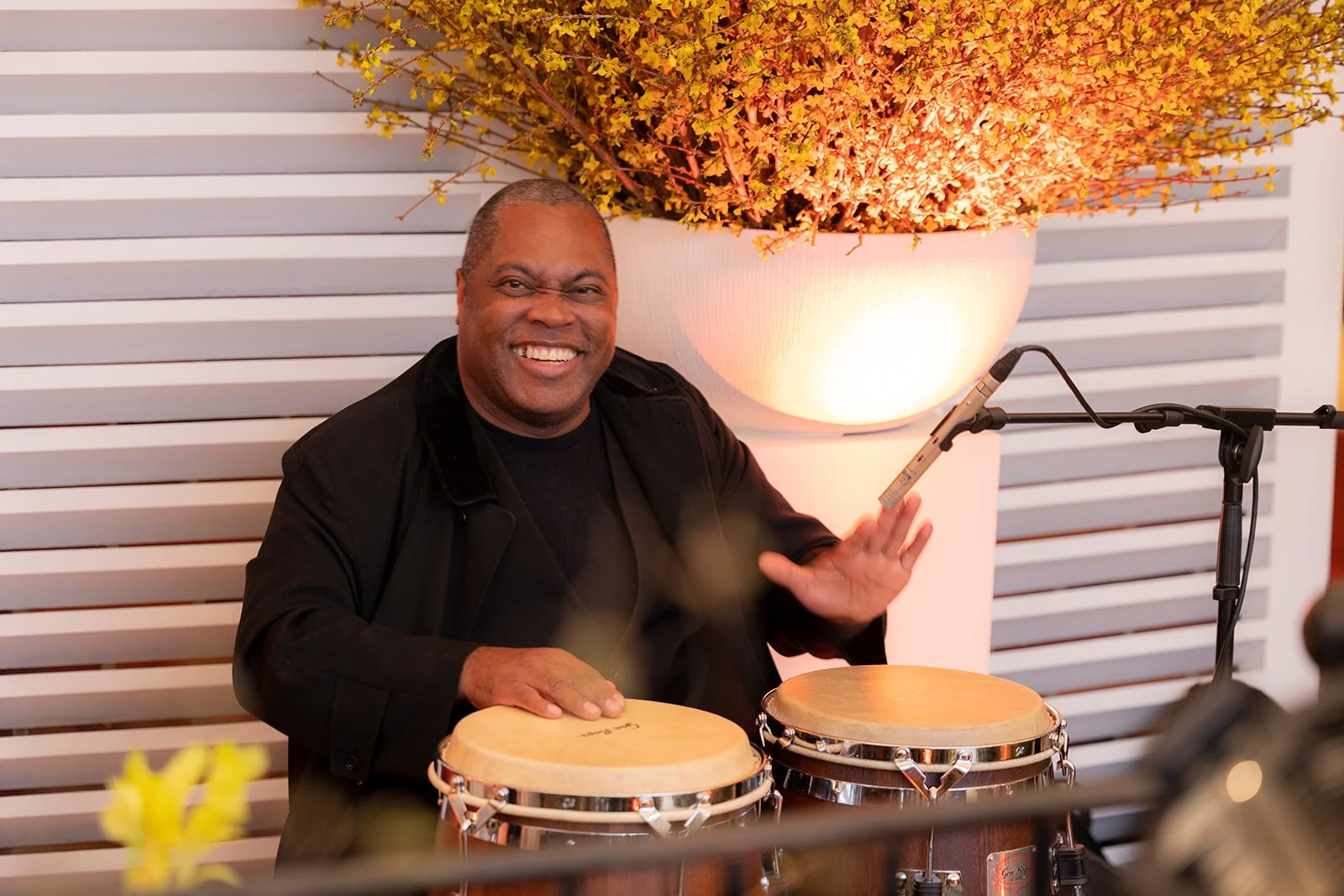A smiling man with short hair playing bongos, seated at a microphone, in an indoor setting with a large decorative plant in the background.