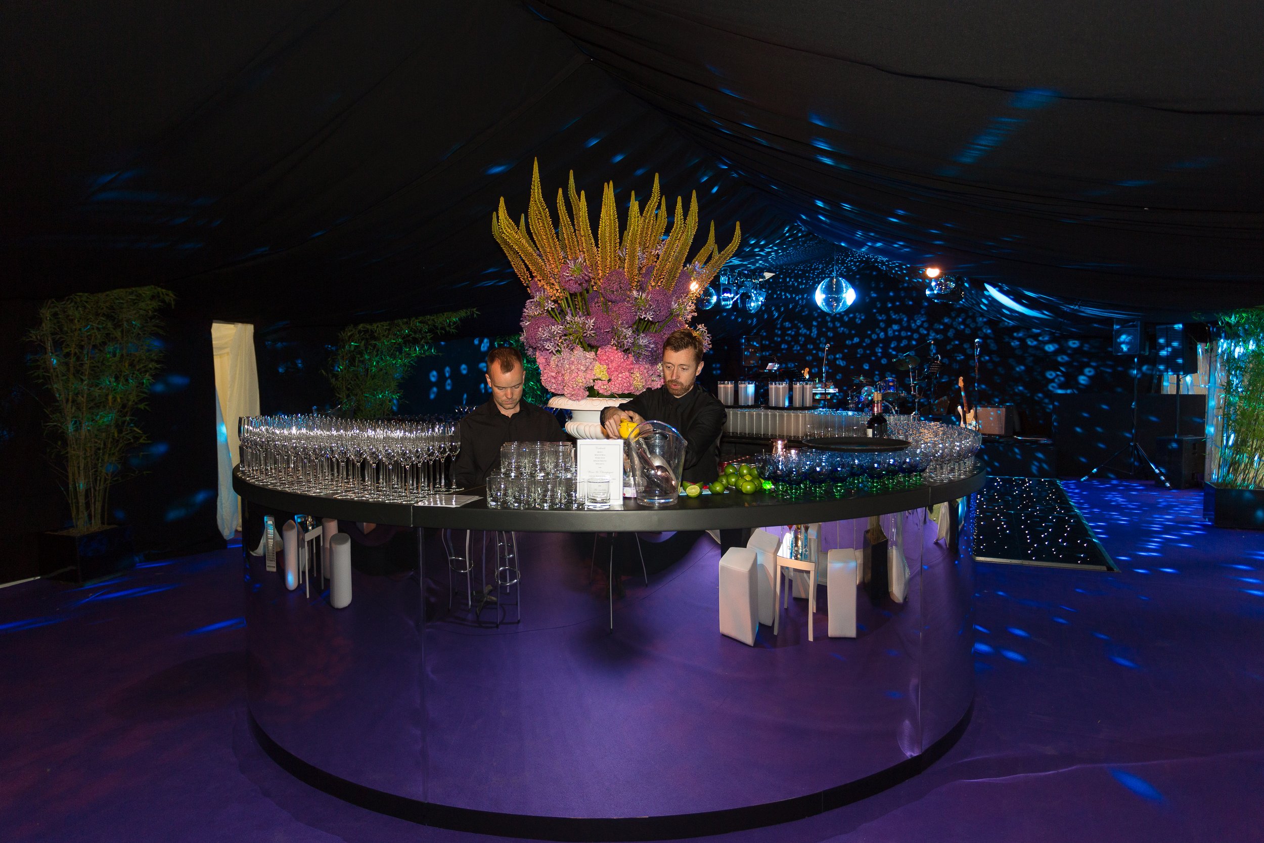 A dark tent decorated with blue lights, featuring a floral arrangement with yellow, pink, and purple flowers on a bar counter where two men are preparing drinks, surrounded by glasses, limes, and bar equipment.