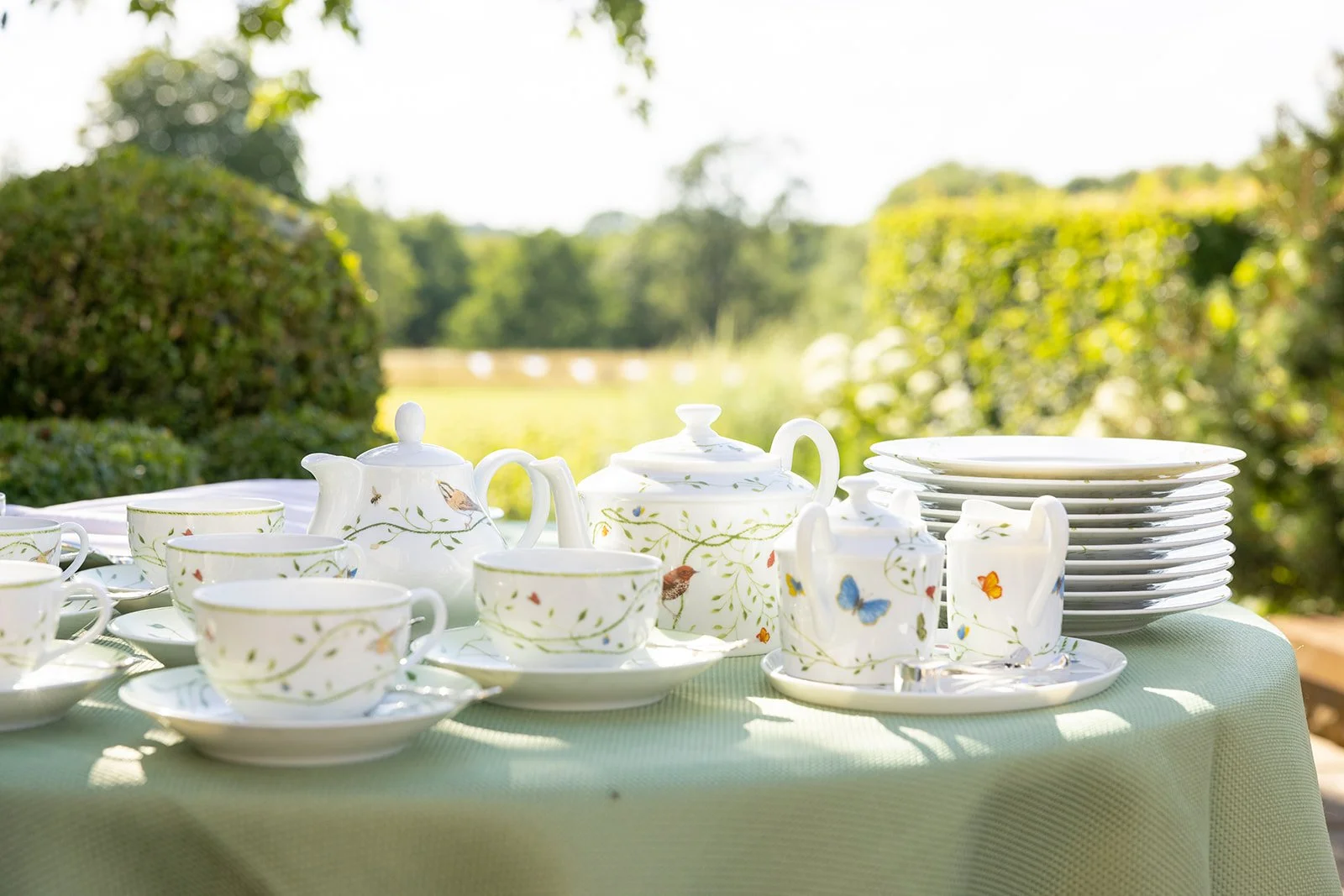 Tea set with butterfly and bird patterns displayed outdoors on a table with a green tablecloth, set against a blurred garden background.