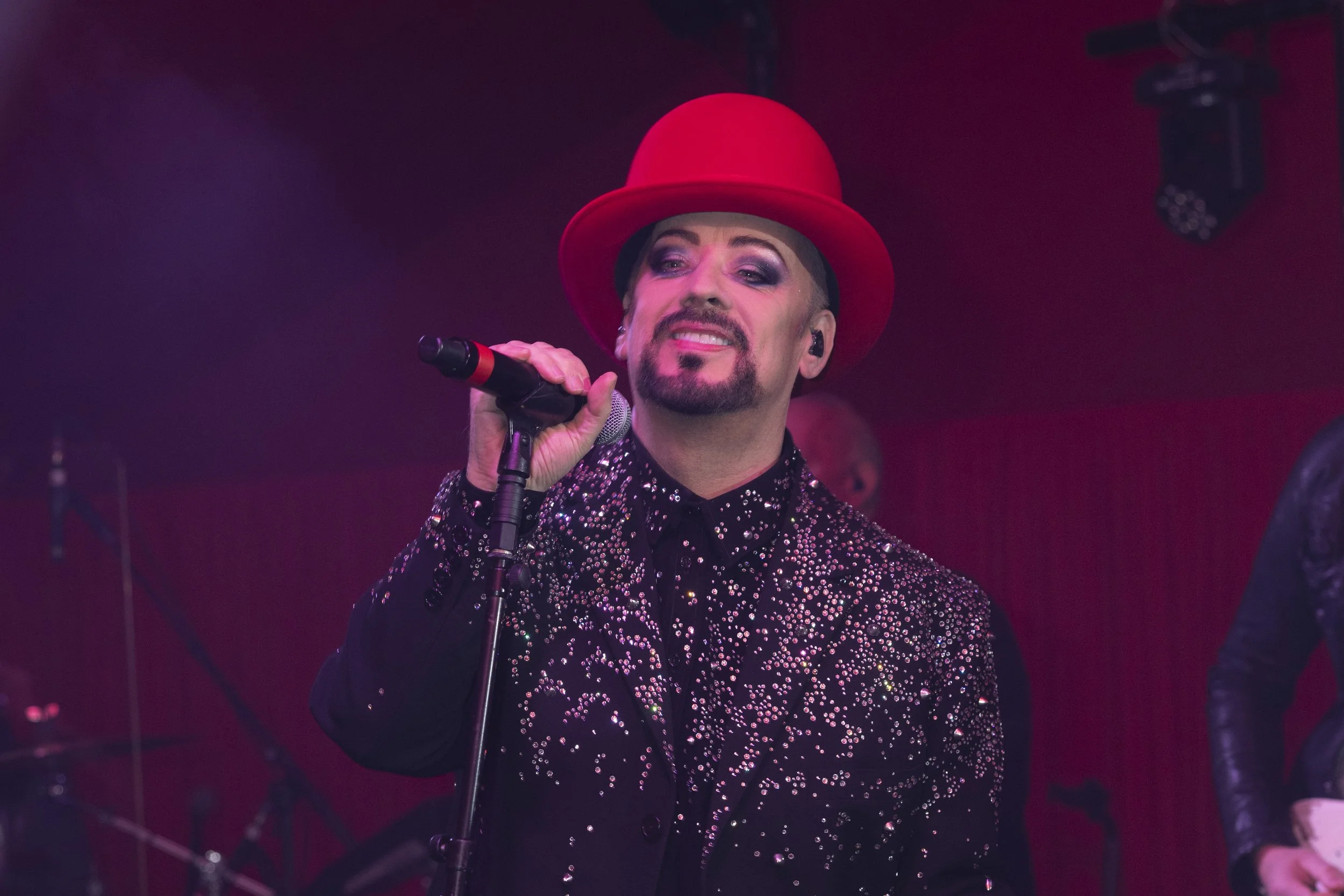 Performer with a red hat, glittery black jacket, and microphone on stage.