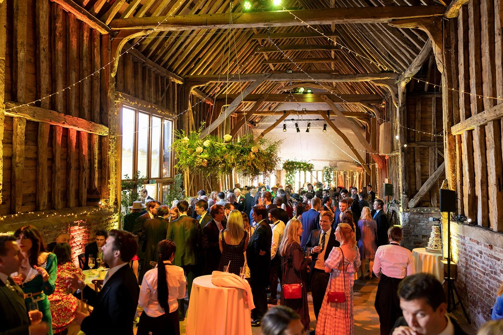 A bustling wedding reception inside a rustic barn with wooden beams, string lights, and floral decorations, filled with guests socializing and standing around cocktail tables.
