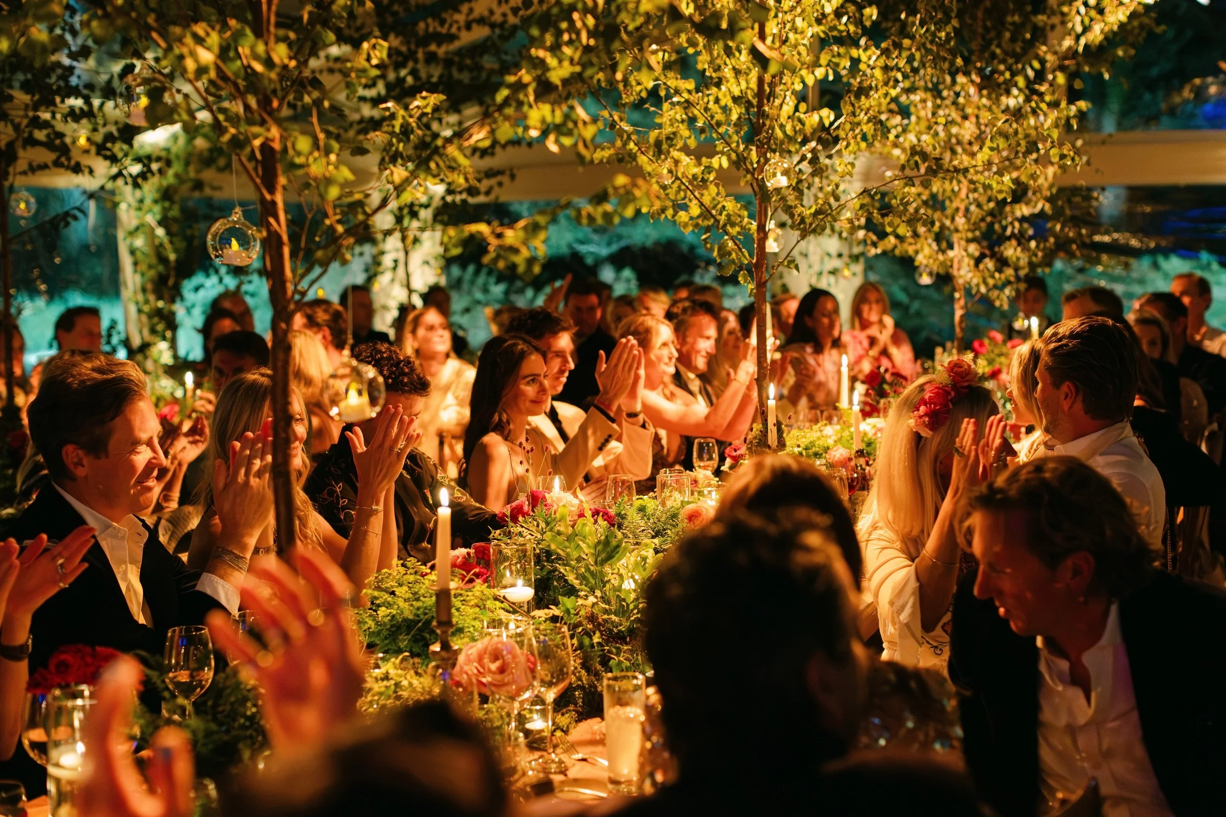 A group of people at a formal dinner party or celebration, surrounded by hanging decorative ornaments and illuminated by candles, in an outdoor setting with trees and greenery.