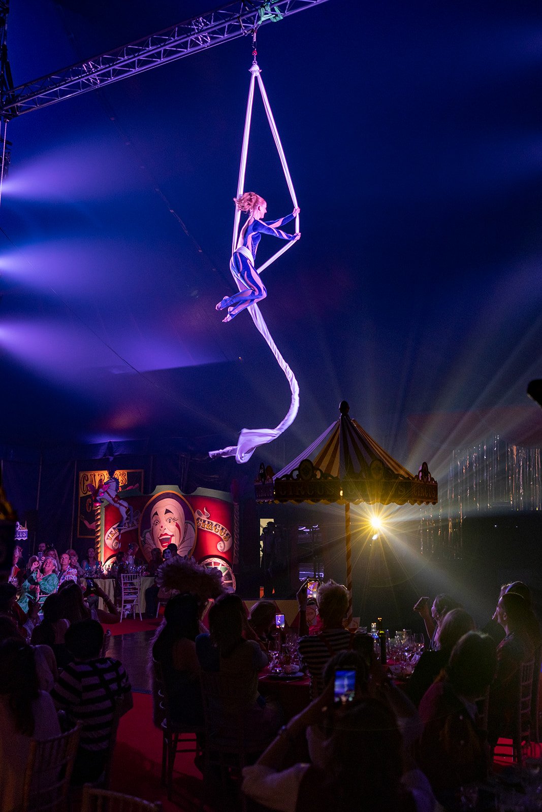 Aerial silk performer in a circus tent, illuminated with purple and blue lighting, with audience seated at tables in the foreground.