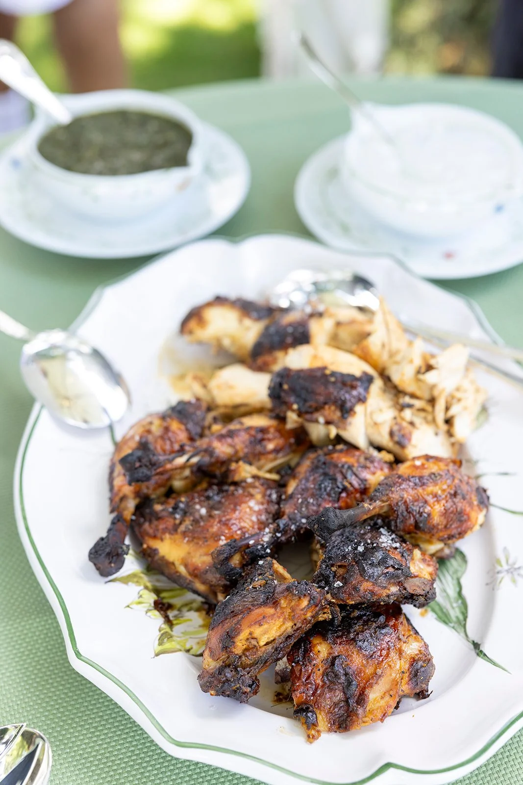 Plate of grilled chicken pieces with charred skin on a decorative white platter, with bowls of sauce or dip and a green tablecloth, outdoors with blurred greenery in the background.