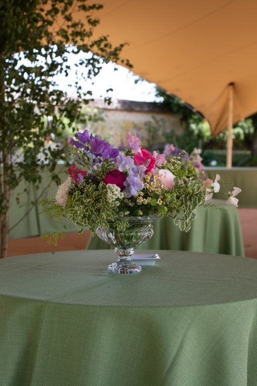 Glass vase filled with colorful flowers on a green-covered round table outdoors under a large tan umbrella.