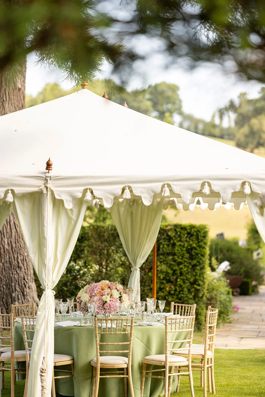 An outdoor event setup with a white canopy tent, decorated with curtains and a floral centerpiece, set in a lush garden area.