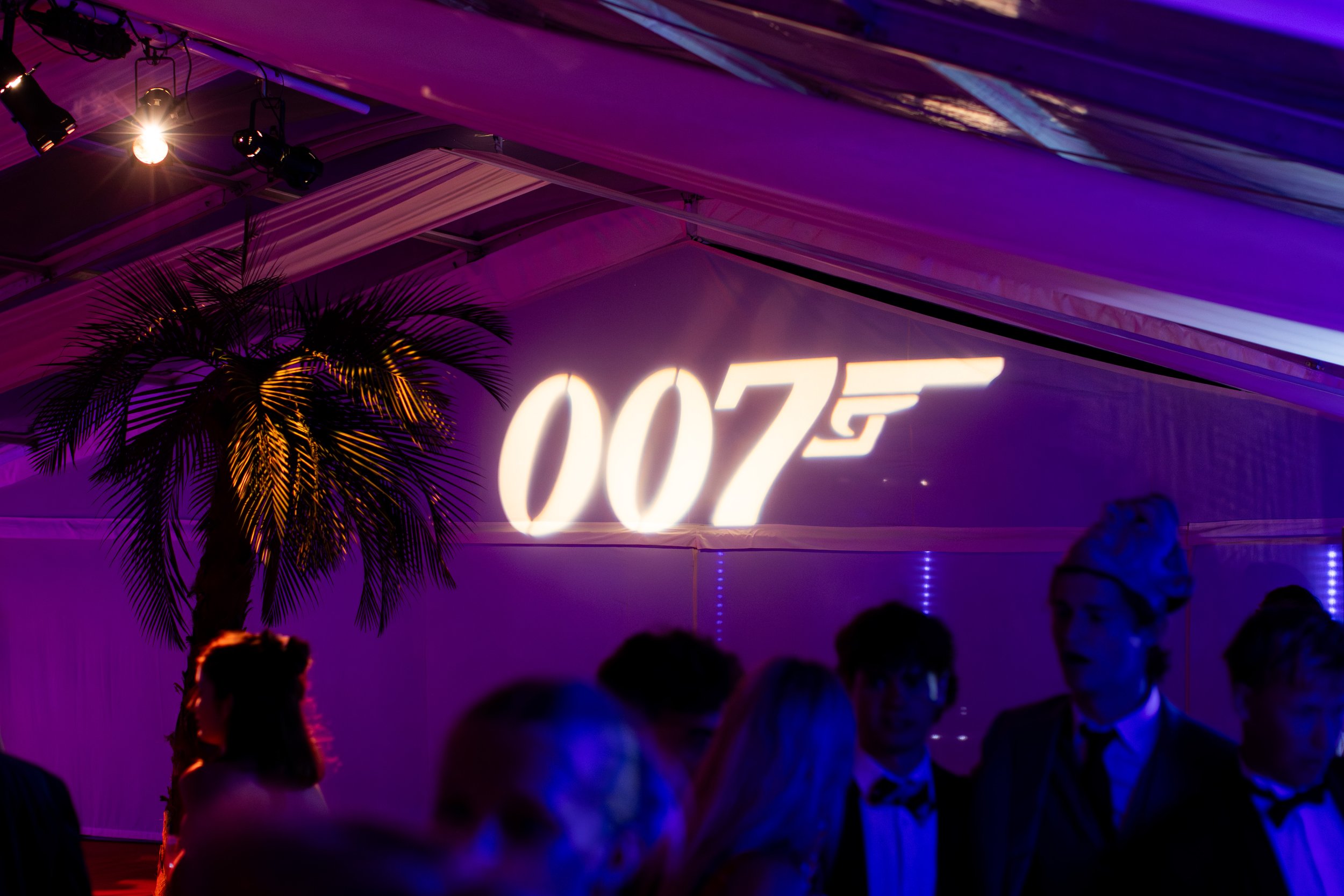 Nightclub with purple and blue lighting, a palm tree silhouette, and a neon sign that reads '007' with people dancing and socializing.