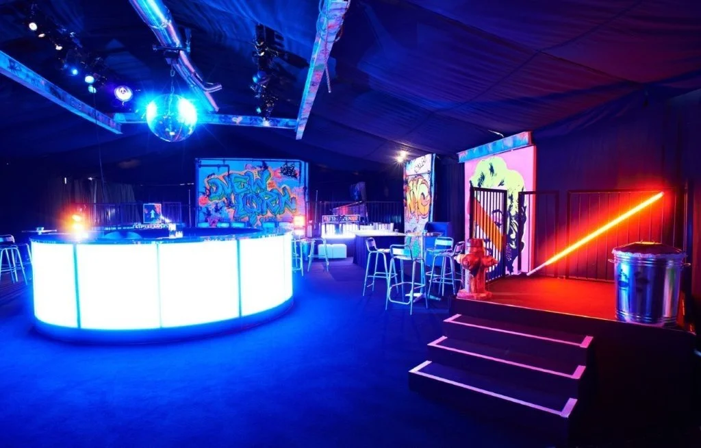 Interior of a nightclub or party space with neon lighting, a bar with glowing blue panels, graffiti artwork on the wall, colorful LED signs, and a small stage area with steps and a trash bin.