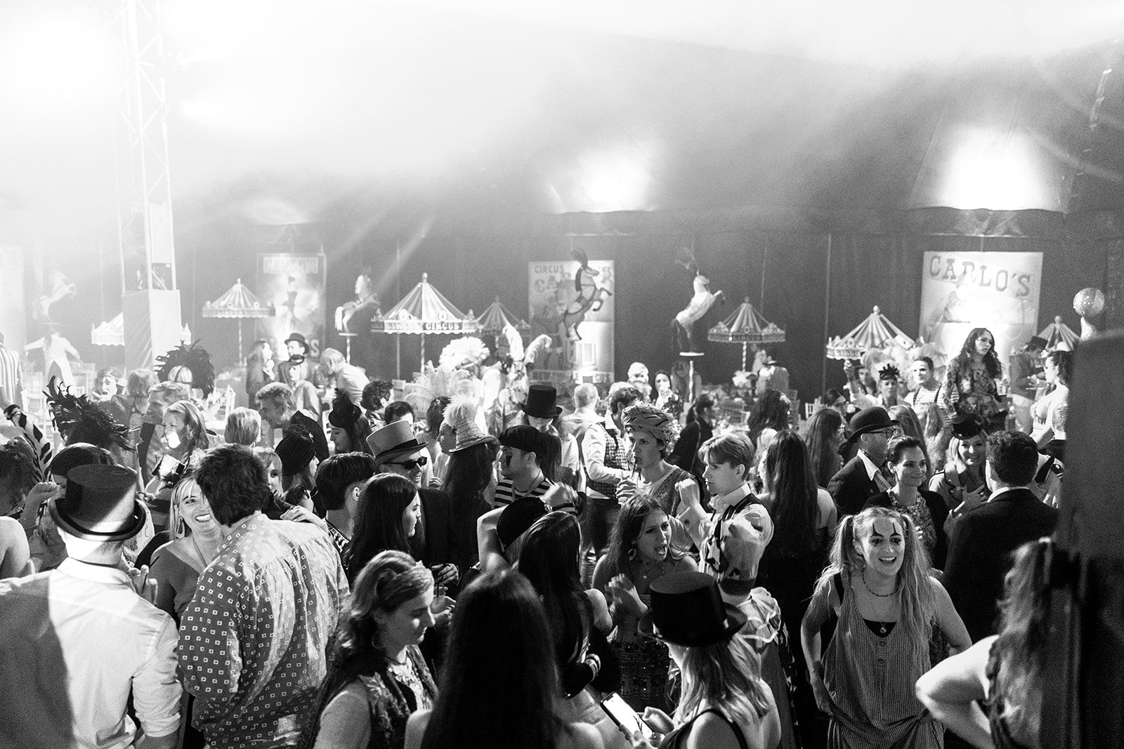 A crowded scene in a circus tent with people dressed in vintage costumes, some with face paint, and some wearing hats and feathered accessories. The background features circus rides and signs, with performers on stilts and a performer riding a horse.