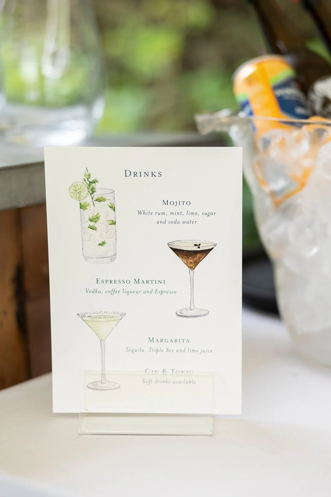 A drink menu with illustrations of a Mojito, Espresso Martini, and Margarita, listing their ingredients, placed on a table with ice buckets and cans in the background.