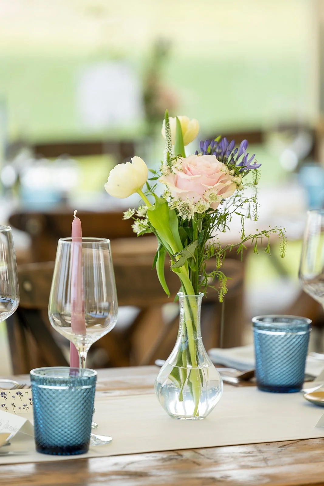 A centerpiece of mixed flowers including pink roses and white tulips in a glass vase on a dining table, with blue glasses and a pink candle in a wine glass nearby.