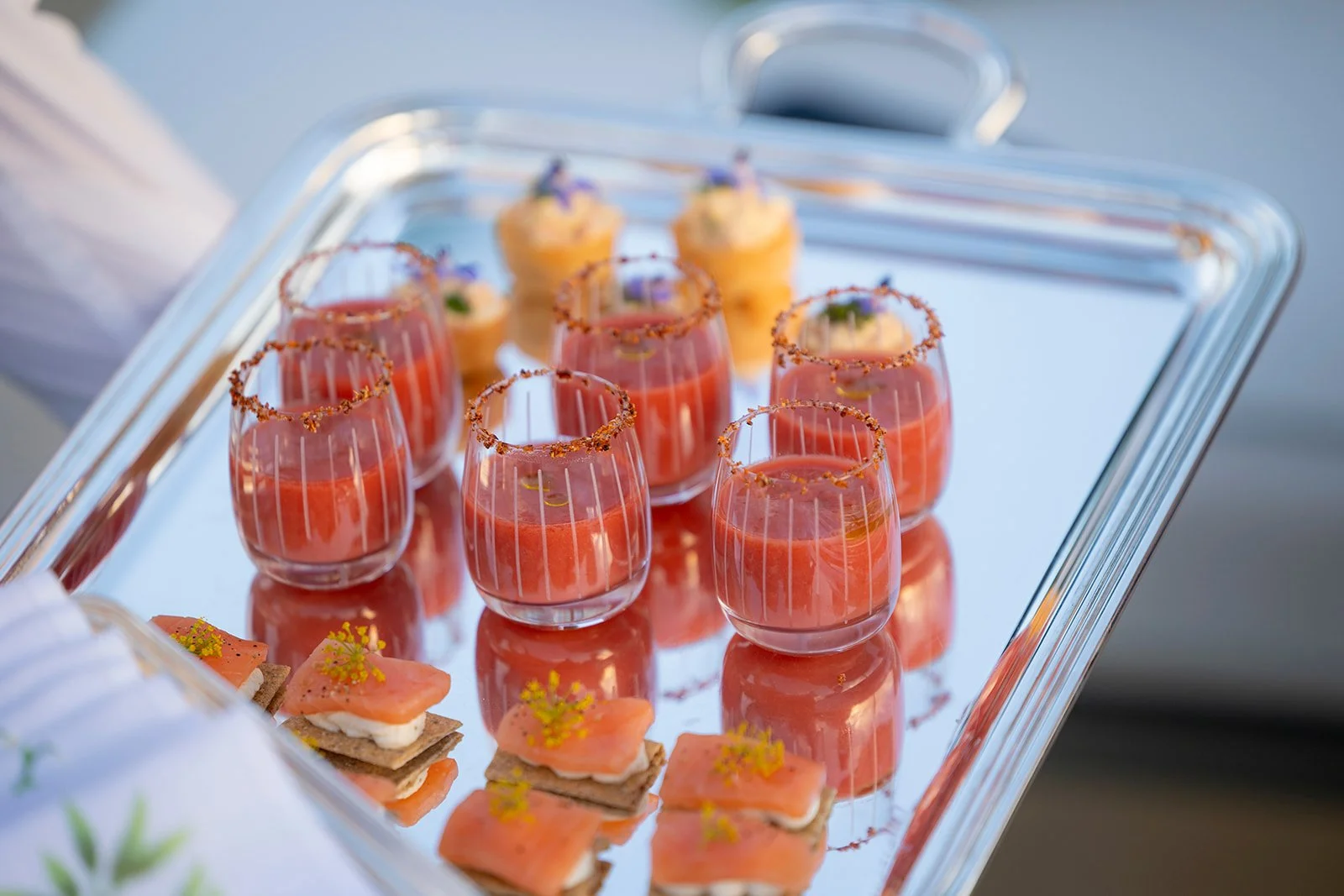 Elegant tray of appetizers including salmon canapés and glasses of pink cocktail with salted rim garnished with herbs.