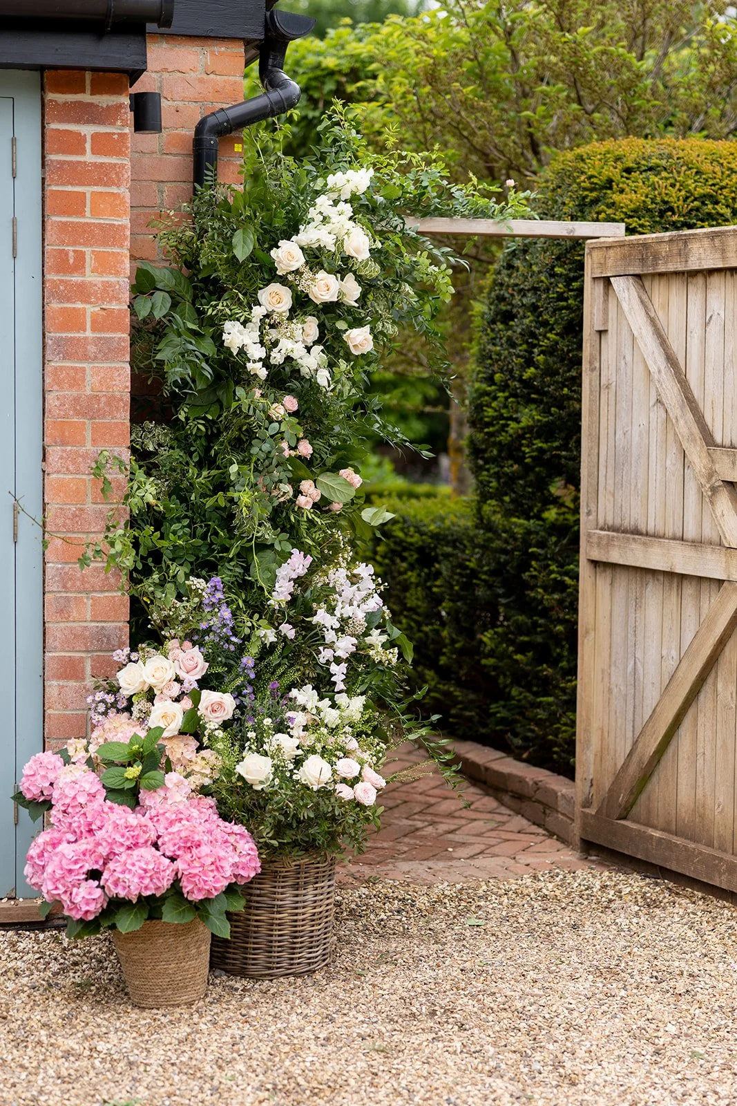 A garden corner with potted pink and white flowers near a deck gate, surrounded by lush green bushes and trees.