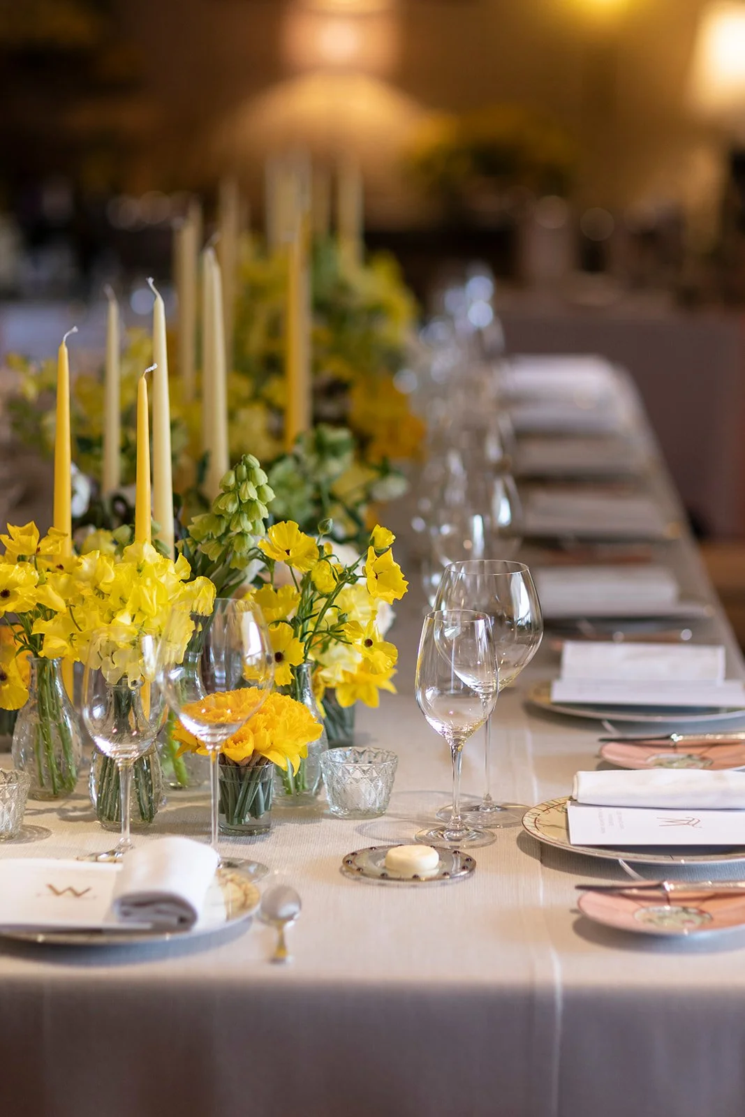 Elegant dining table decorated with yellow flowers, candles, and glassware, set for a formal event.
