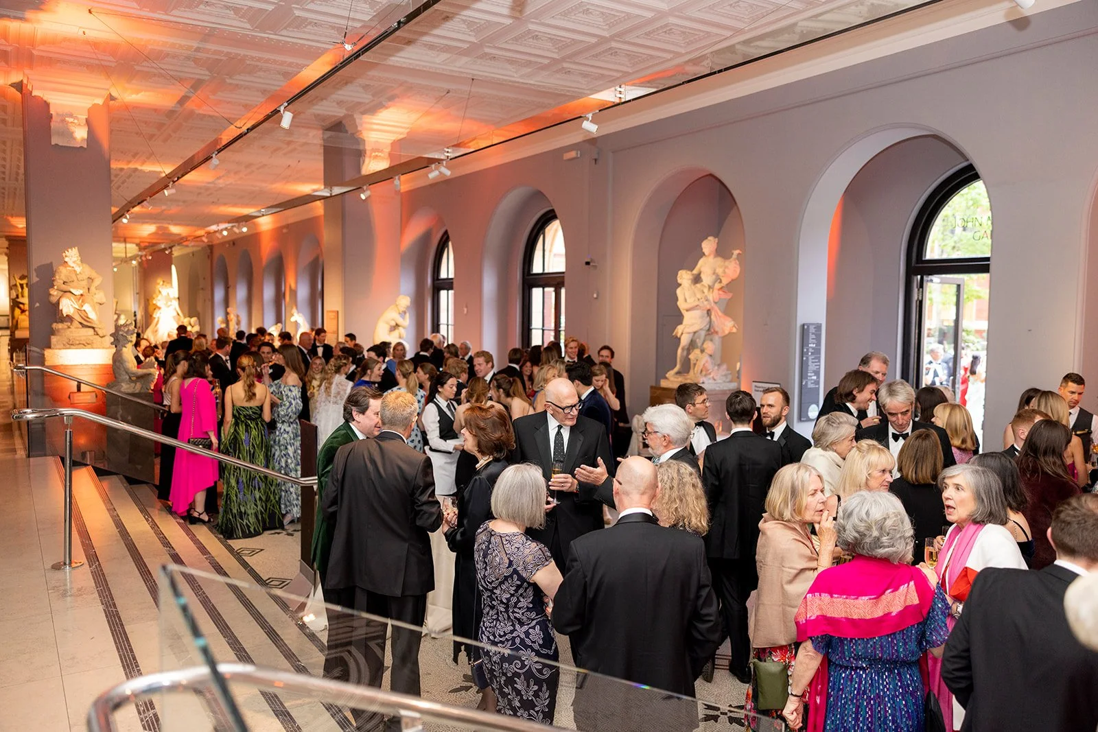 Elegant ballroom filled with well-dressed people socializing at a formal event, with classical sculptures and large arched windows along the walls.