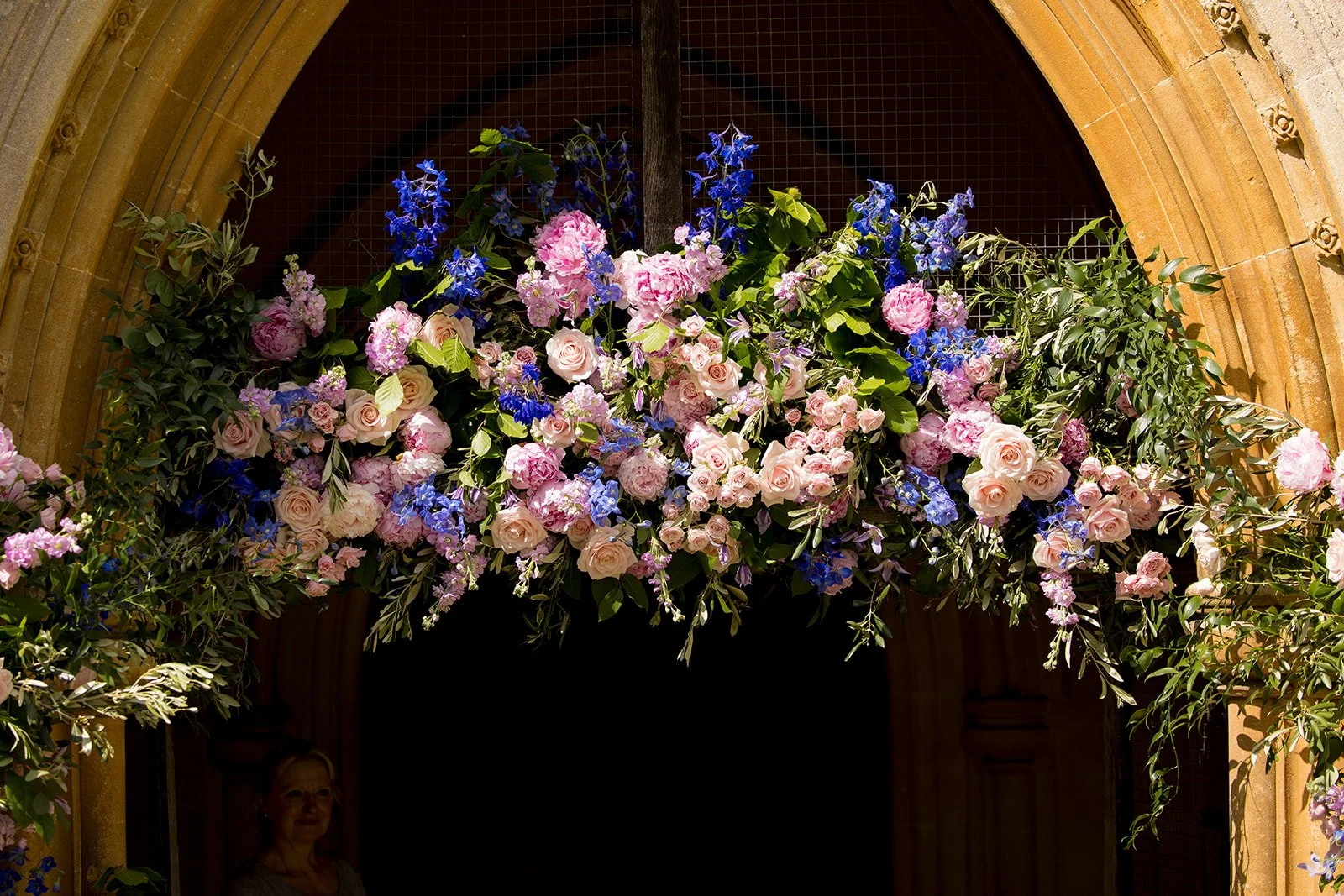 A floral arrangement of pink and blue flowers with green leaves hanging above an archway in a church or cathedral interior.