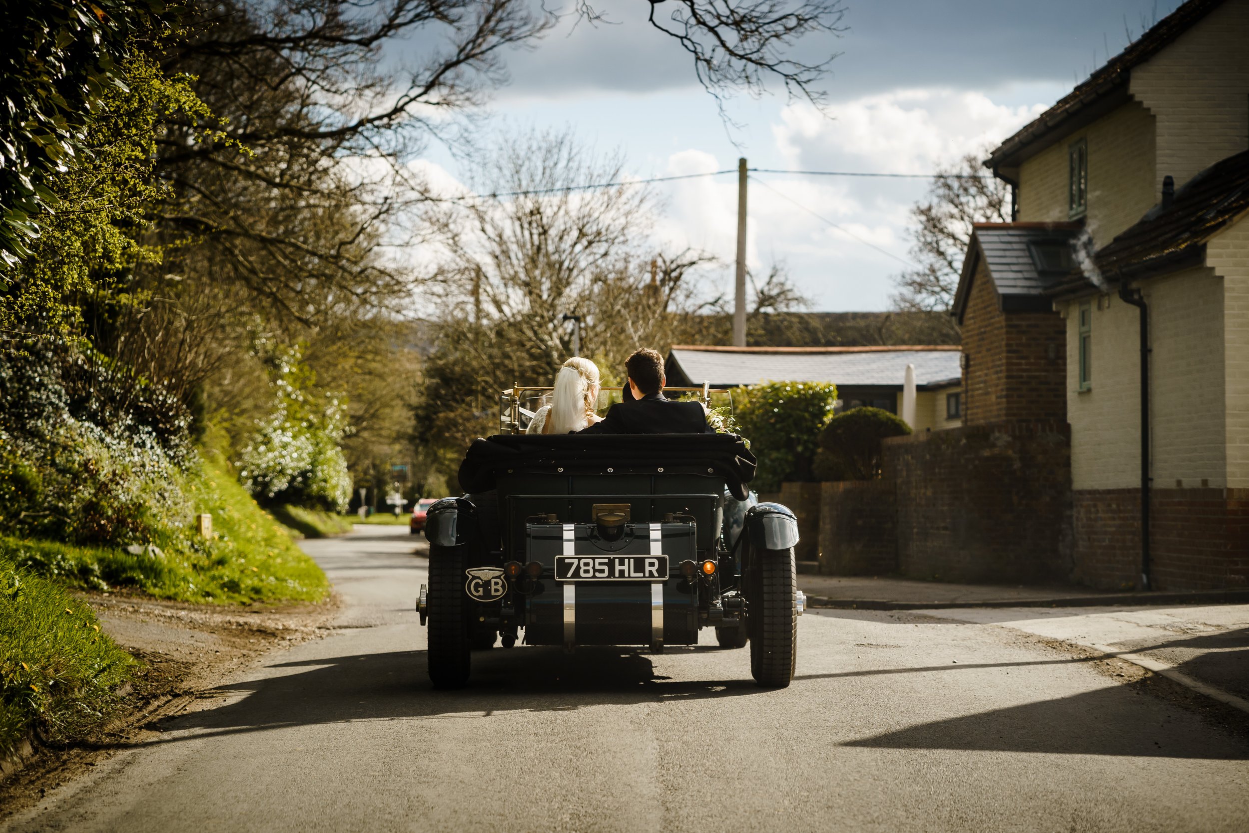 A vintage black car with a license plate reading '785 HLR' and a 'GB' sticker on the bumper is being driven down a residential street. Two people, a woman with a white headscarf and a man, are sitting in the car. The street is lined with trees, some 