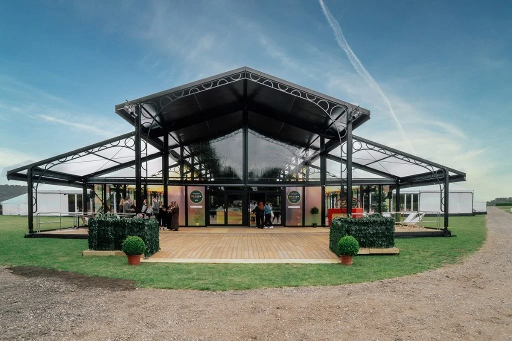 A large, modern event tent with glass walls and a black metal frame, set up outdoors on a grassy field, with people inside and a clear blue sky overhead.