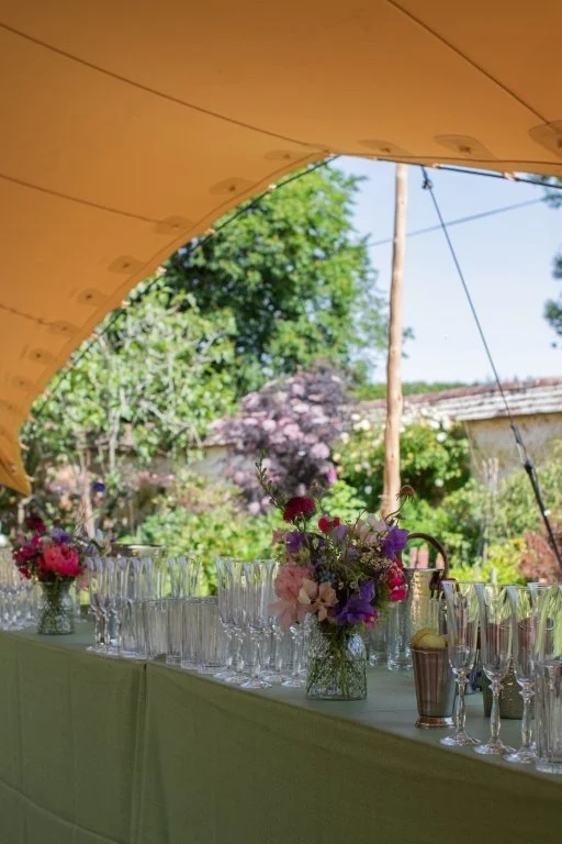 A long table set outdoors under a canopy with flowers and glassware, with greenery and houses in the background.