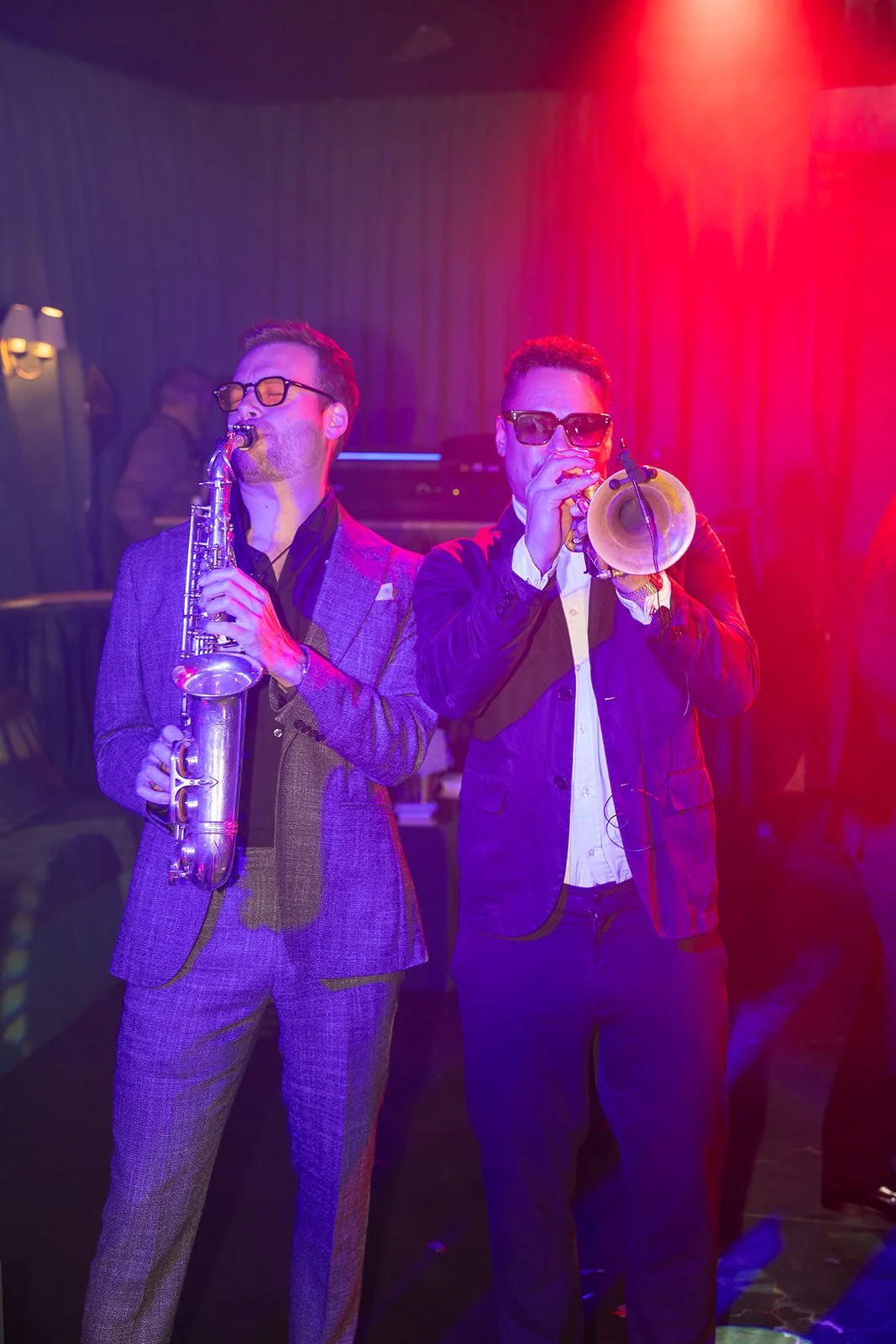 Two men performing jazz music, one playing saxophone and the other playing trumpet, under colorful stage lighting.
