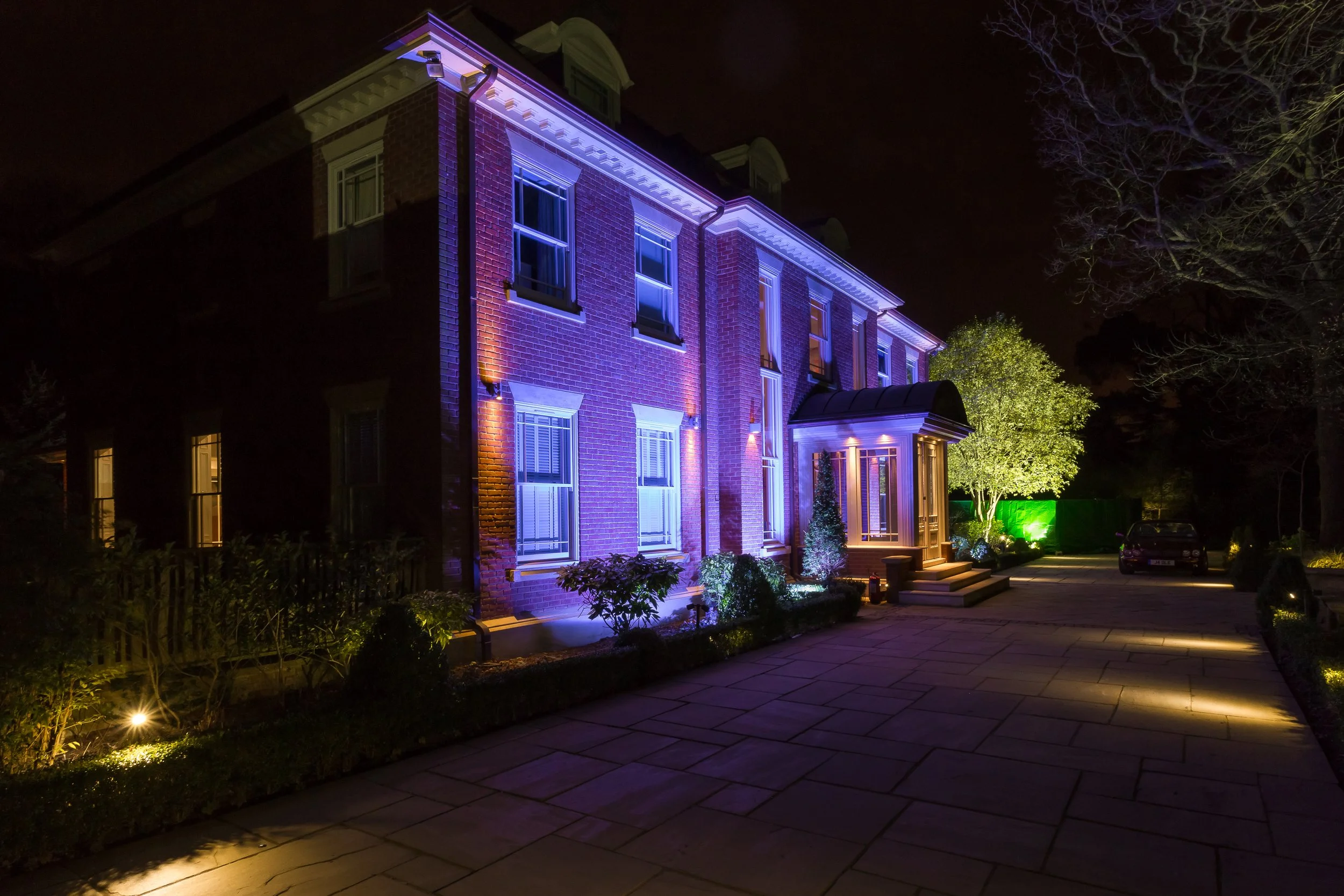 Nighttime view of a brick building illuminated with colorful lights, with trees and a paved walkway in the foreground.