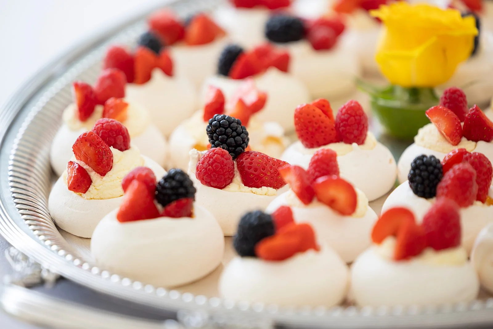 Mini pavlova desserts topped with fresh strawberries, raspberries, and blackberries on a silver tray with a yellow flower in the background.