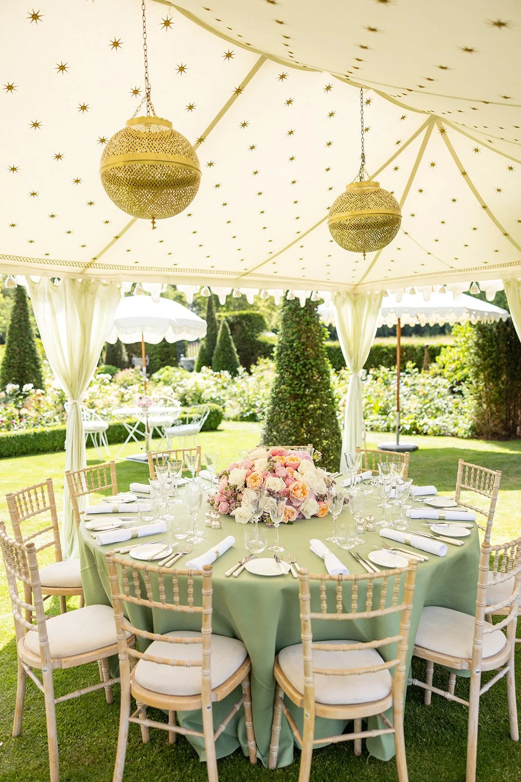 Elegant outdoor dining setup under a white canopy with gold star decorations, featuring a round table with a floral centerpiece, surrounded by wooden chairs with white cushions, set with dishes and glassware, with a lush garden in the background.