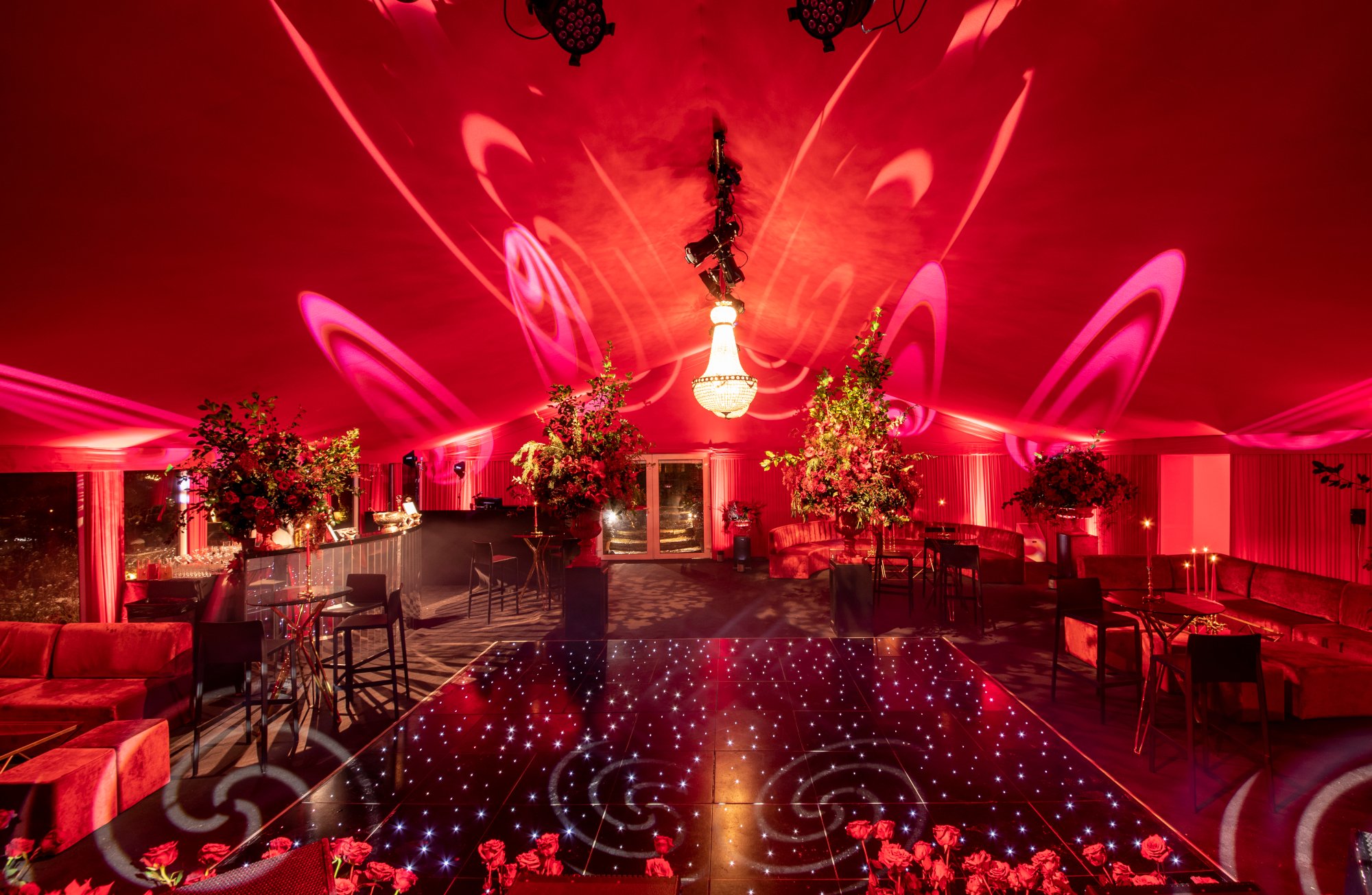 A lavish event space illuminated with pink and red lighting. The ceiling is decorated with a chandelier and spotlights, creating swirling light patterns. The room features floral arrangements and tables for mingling, with a dance floor decorated with star-like lights at the center.