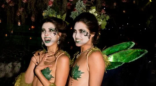 Two women dressed as fairies with green makeup, green leaf pasties, and butterfly-like wings, standing outdoors at night with decorative lighting.