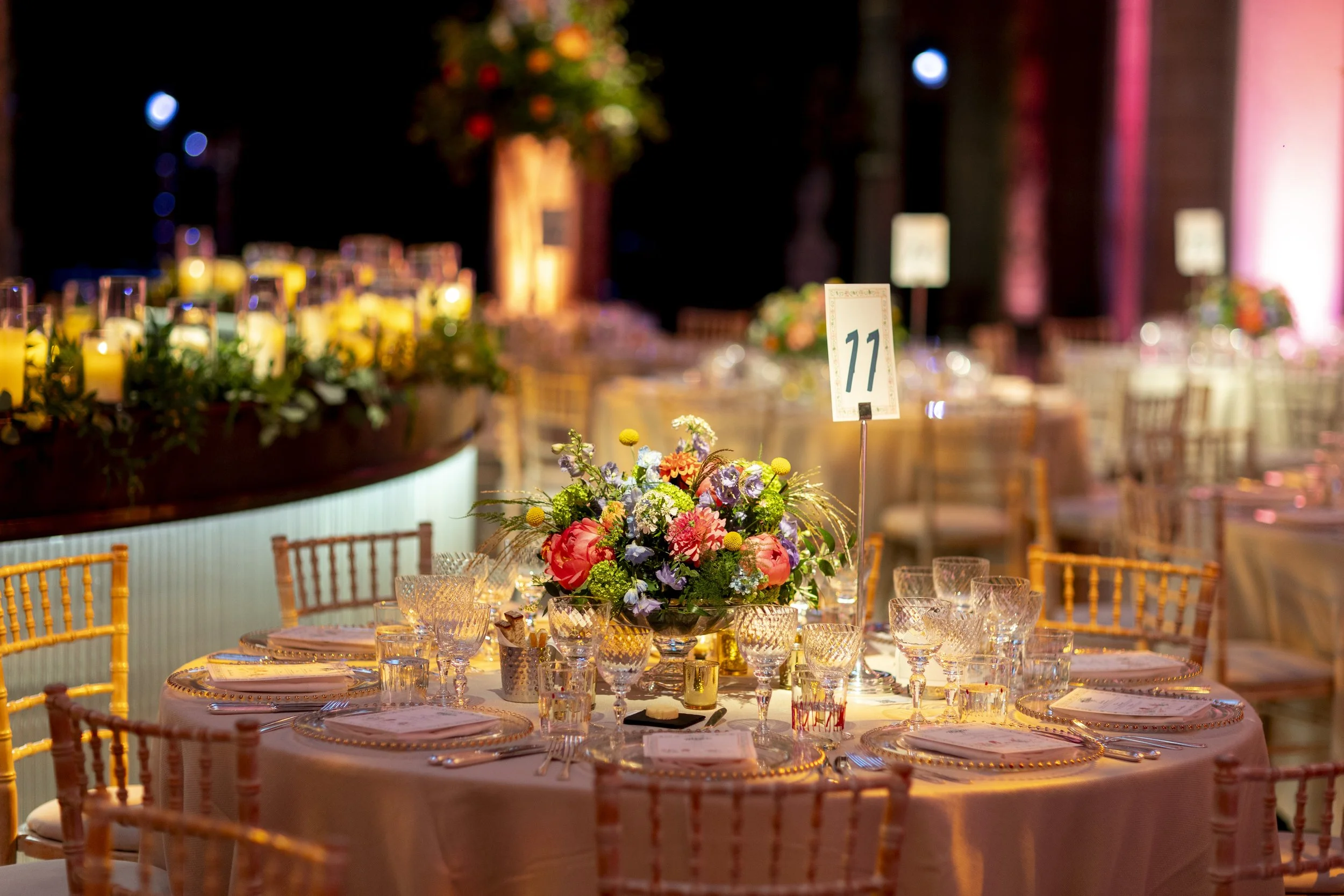 A decorated banquet hall at night with a pink and purple color theme, featuring a long dining table with a floral centerpiece, glassware, and place settings, and a table number card with the number 11.