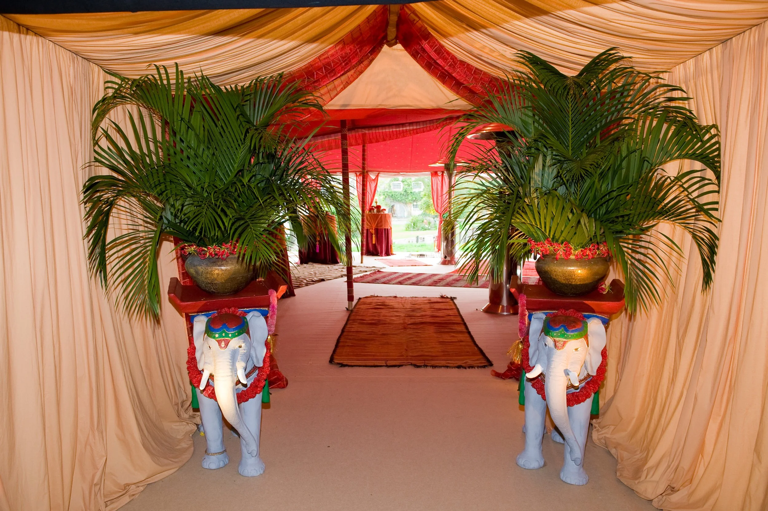 Decorated entrance with two white elephant statues adorned with red garlands and colorful painted details, flanking a pathway leading to a red canopy in an event tent decorated with beige drapes and red accents, set outdoors.