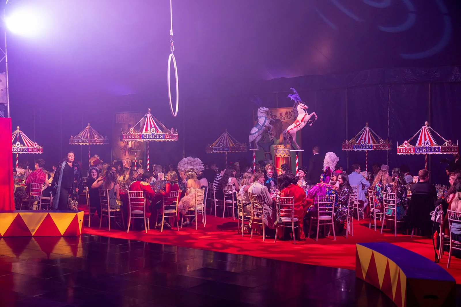 A lively circus-themed dinner event with children and adults seated at round tables, decorated with classic circus tents and statutes of horses on stilts, under purple lighting.