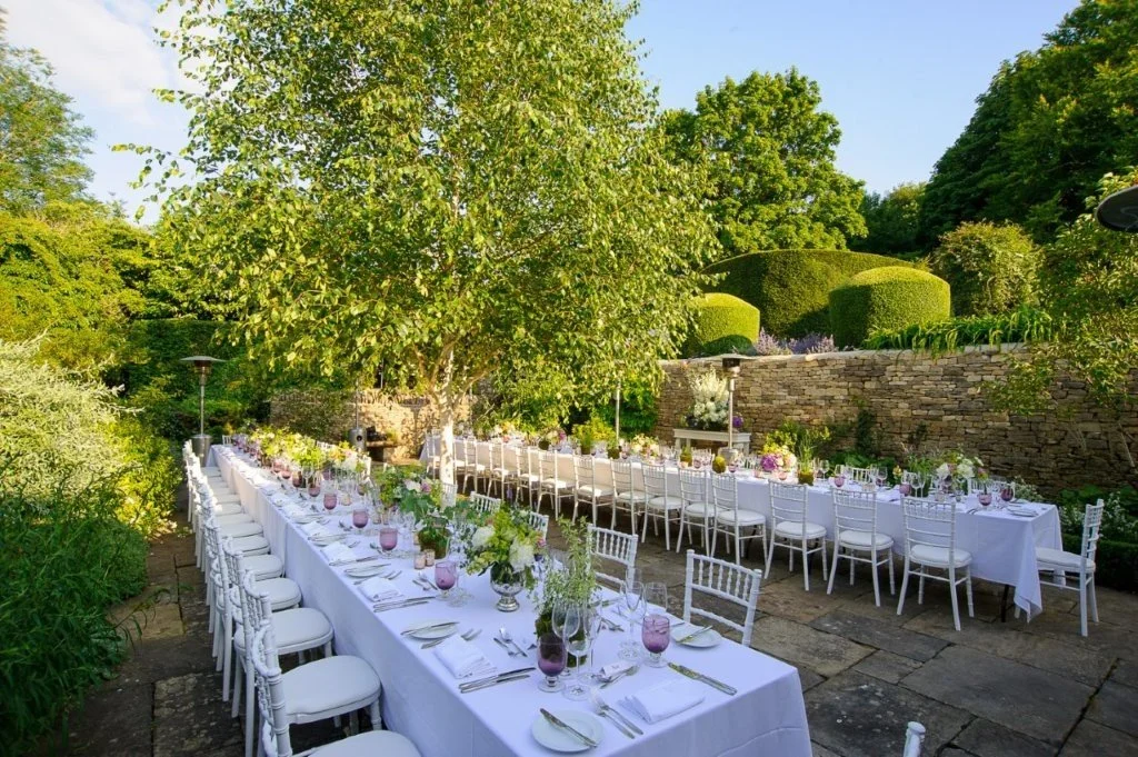 An outdoor garden dining setup with long white-clothed tables, white chairs, and purple glassware, set in a lush green garden with trees, shrubs, and a stone wall.