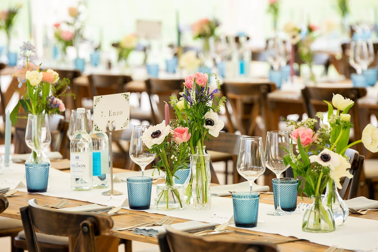 Elegant banquet table set with colorful floral centerpieces, wine glasses, water bottles, and place settings in a bright, airy room for a wedding or event.