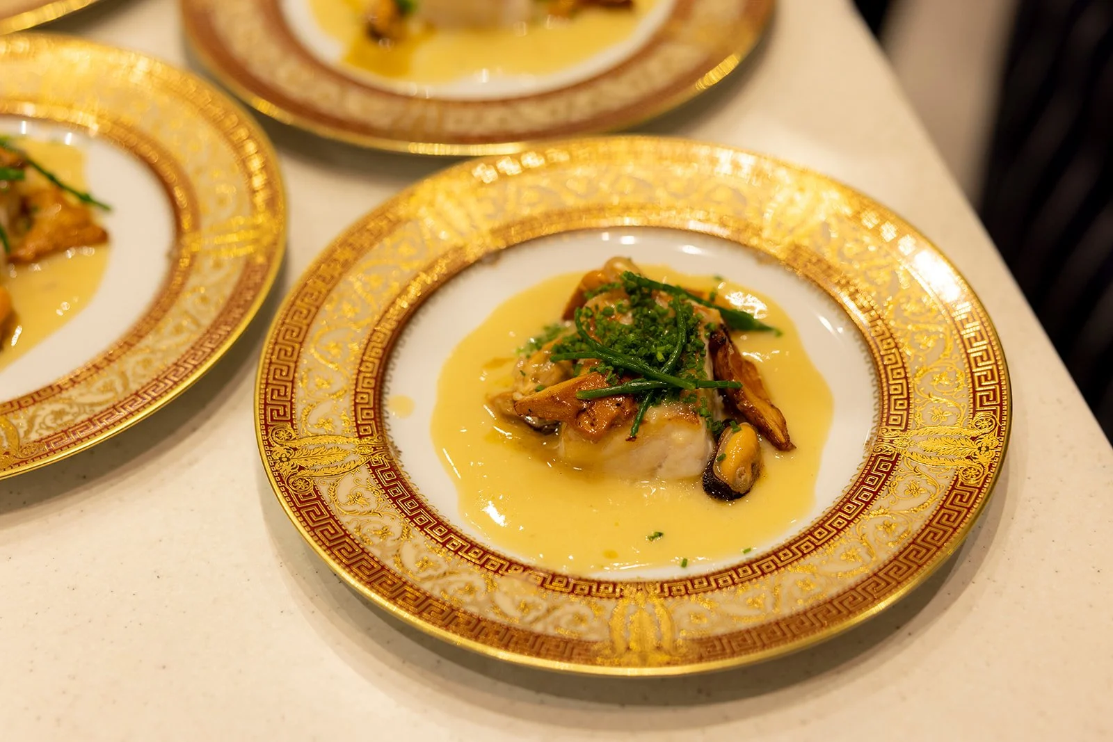 Dish with fish topped with green herbs and mushrooms, served in a creamy yellow sauce on an ornate gold and white plate.