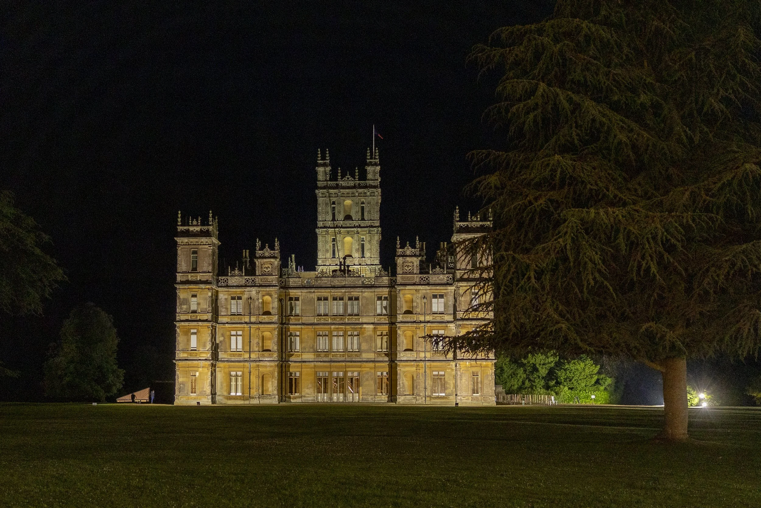 Night view of a historic castle illuminated, surrounded by green trees, with a dark sky overhead.