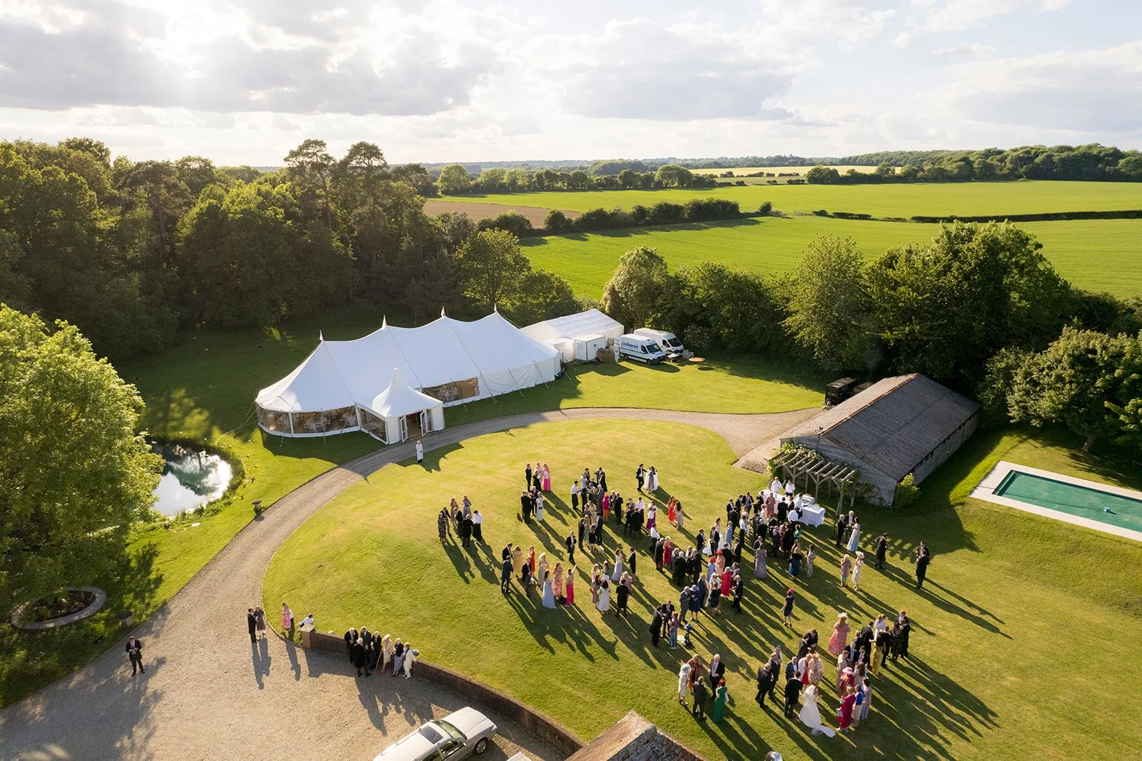 Outdoor wedding reception in a lush green field with a large white tent, guests dressed in formal attire, and scenic countryside landscape in the background.